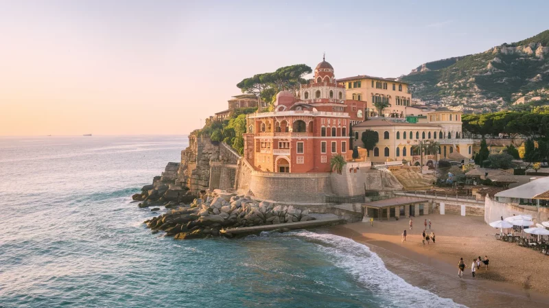 Una città costiera con edifici color pastello adagiati su una costa rocciosa, caratterizzata da una piccola spiaggia sabbiosa e un mare calmo sotto un cielo mite al tramonto.