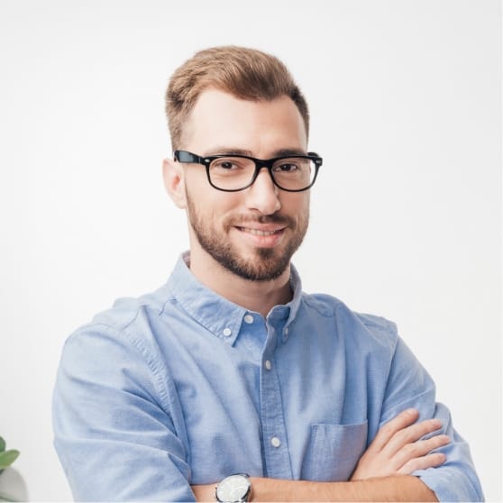 Un uomo con barba e occhiali, che indossa una camicia azzurra abbottonata, è in piedi con le braccia incrociate e guarda la telecamera su uno sfondo bianco.