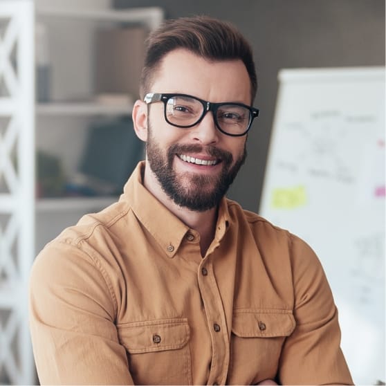 Un uomo con barba e occhiali, che indossa una camicia marrone, sorride in un ufficio davanti a una lavagna.